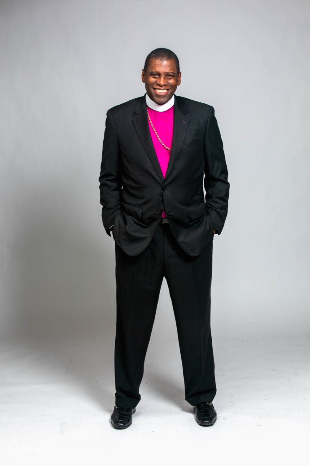 Bishop Douglas standing, smiling, in a black suit and fuchsia clerical shirt